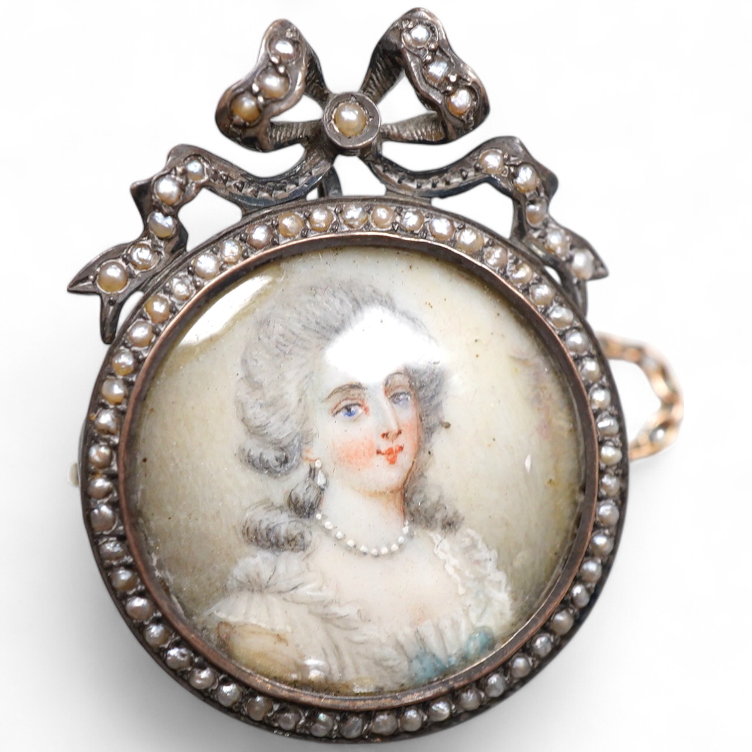 A painted miniature and seed pearl brooch, late 19th century, set with a glazed circular portrait miniature on ivory depicting a lady dressed in the 18th century manner, within a mount topped with a ribbon bow, set with